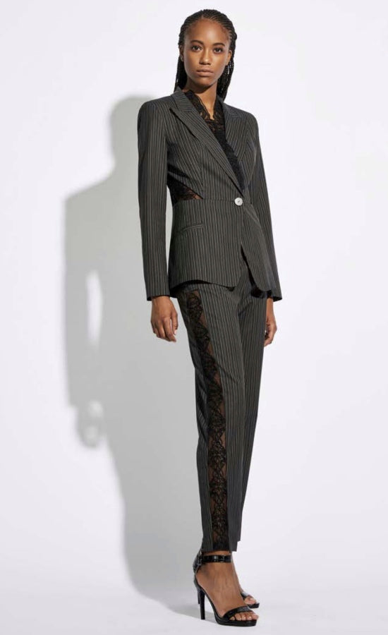 Front full body view of a woman wearing a black and white striped blazer with a single button and mesh detailing. On the bottom she is wearing the Oblique Creations Striped Trouser. This trouser is also striped black and white to match the blazer. On the right leg there is a detailed black mesh see-through band running down the side of the leg. 