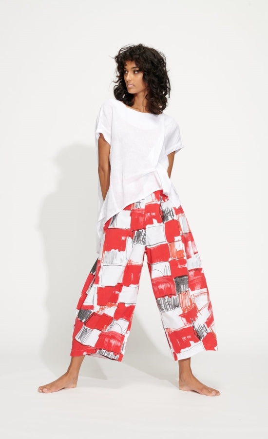 Front full body view of a woman wearing a white top and the banana blue red patchwork pant. This pant is wide legged and ends above the ankles. It is white with red and black squares painted all over it.