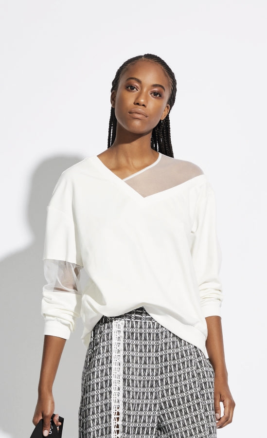 Front, top half view of a woman wearing black and white printed pants with the Oblique Creations Sweatshirt on top. The sweatshirt is white with mesh detailing on the left side of the neck line and in the middle of the long sleeves. The v-neck is off center.