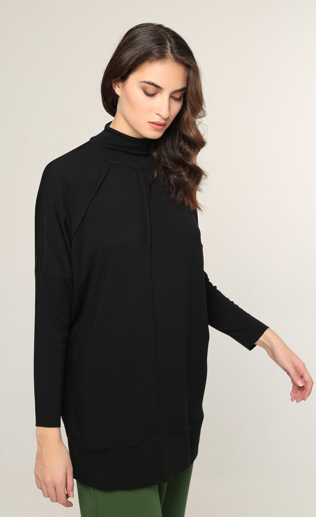Front top half view of a woman wearing the ozai n ku black top. This top is solid black with long sleeves and drop shoulders. The top sits below the hips and features decorative seams and a high neck.