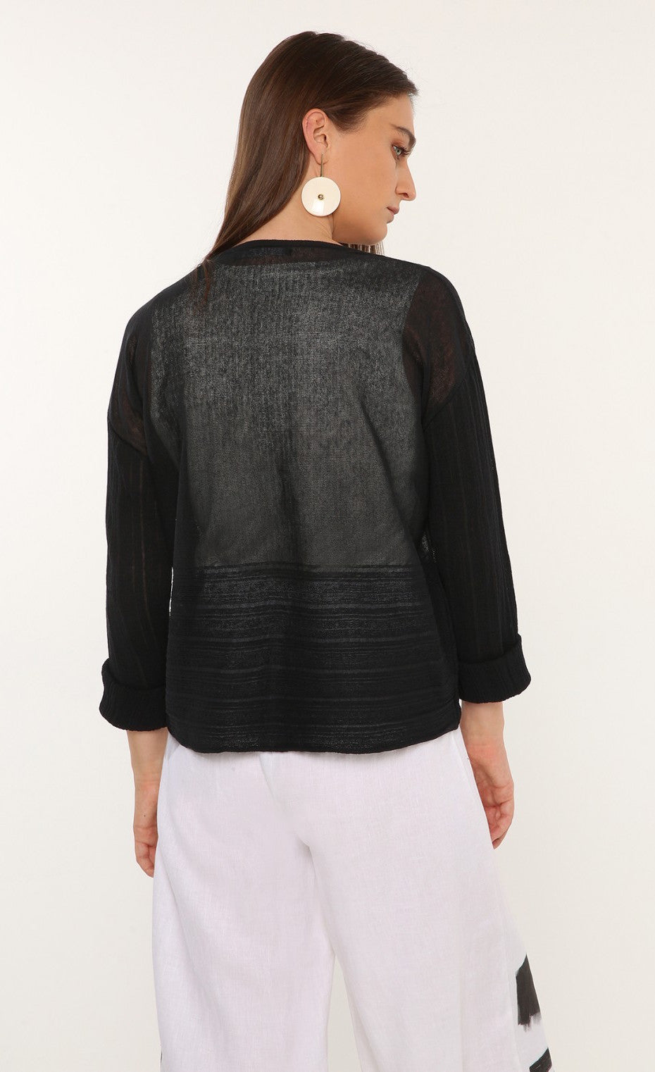Back top half view of a woman wearing the ozai n ku mimosa knit top. This top is black and translucent. It has a darker pattern running across the bottom of the back and on the long sleeves. 