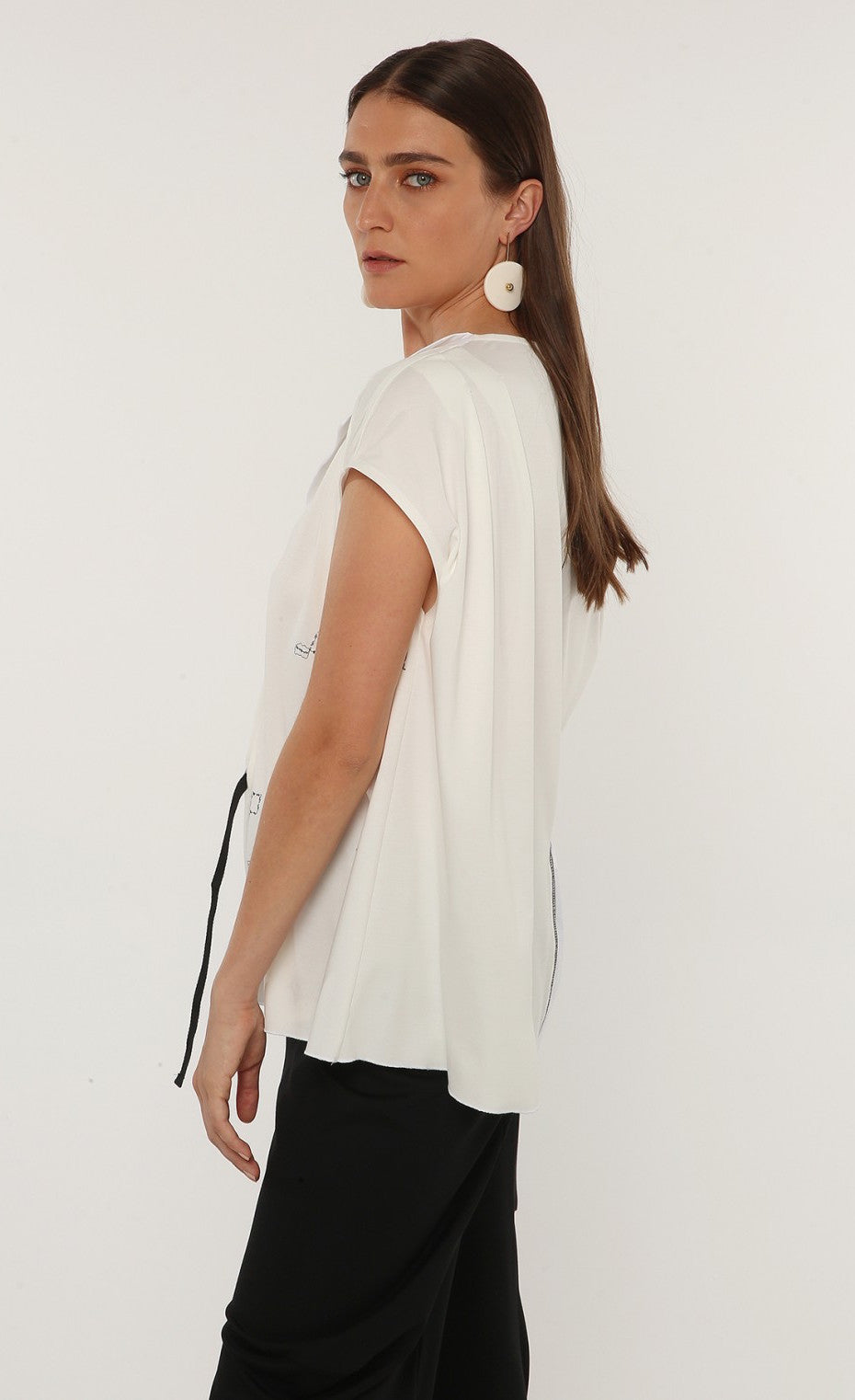 Left sided top half view of a woman wearing the ozai n ku tuberose top. This top is white and off-white. The front has cap sleeves, black little sketched designs, a split v-neck, and a drawstring waist on the right side.