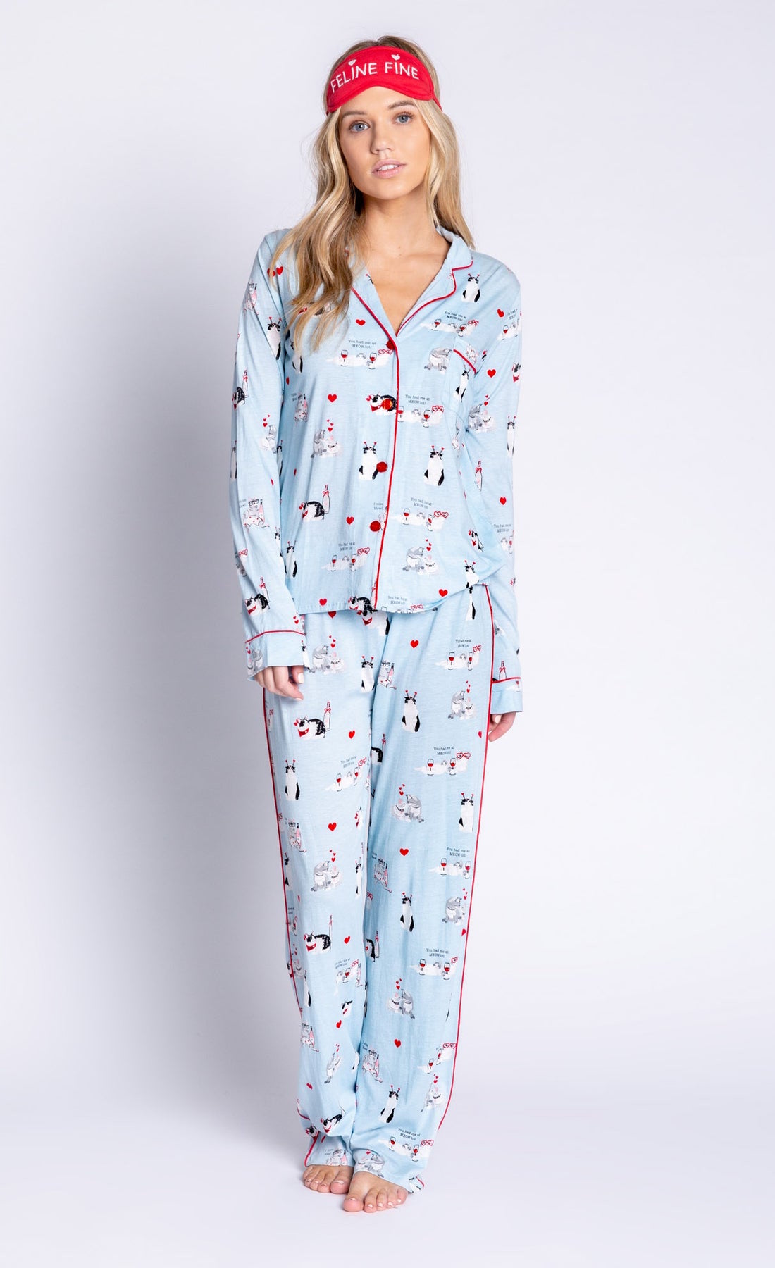 Front full body view of woman wearing the pj salvage love is a four legged word pj set. This set is light blue colored with cats printed all over it and red trim. The set has a button down shirt with long sleeves, relaxed pants, and a red sleep mask that says feline fine.