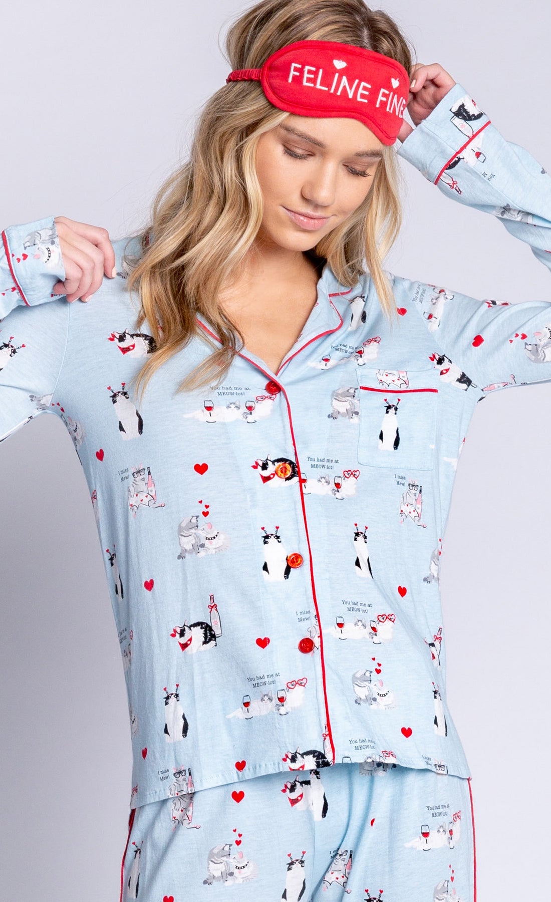 Front top half view of woman wearing the pj salvage love is a four legged word pj set. This set is light blue colored with cats printed all over it and red trim. The set has a button down shirt with long sleeves and a red sleep mask that says feline fine.