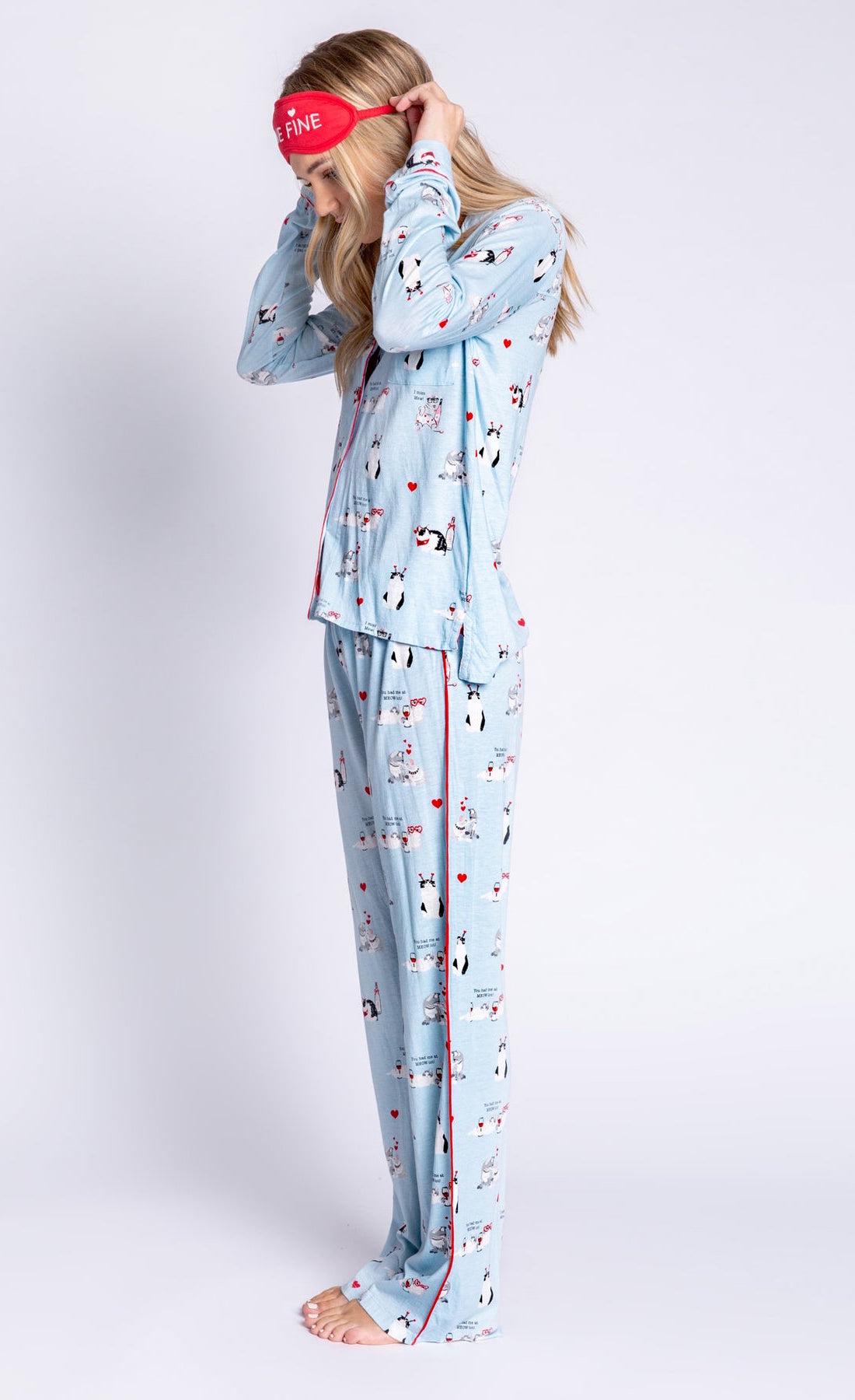 Left side full body view of woman wearing the pj salvage love is a four legged word pj set. This set is light blue colored with cats printed all over it and red trim. The set has a button down shirt with long sleeves, relaxed pants, and a red sleep mask that says feline fine.