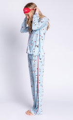 Load image into Gallery viewer, Left side full body view of woman wearing the pj salvage love is a four legged word pj set. This set is light blue colored with cats printed all over it and red trim. The set has a button down shirt with long sleeves, relaxed pants, and a red sleep mask that says feline fine.
