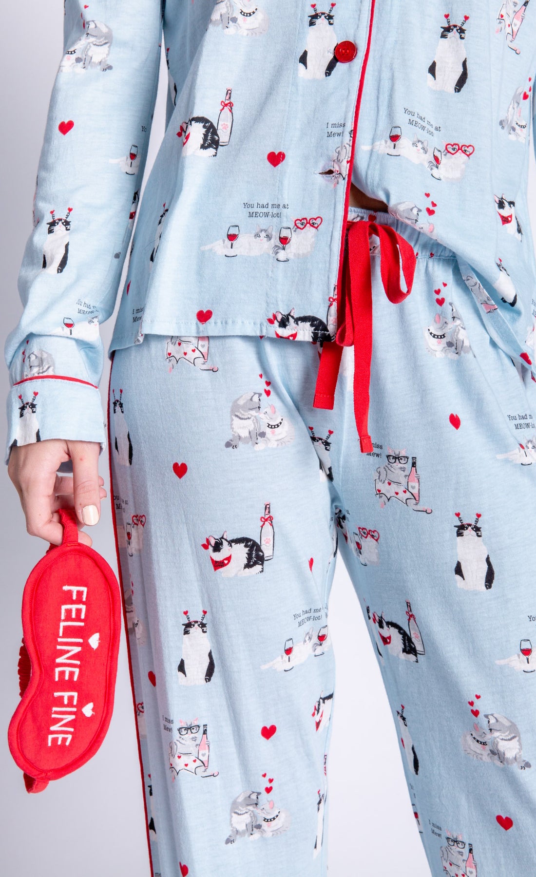Front close up view of woman wearing the pj salvage love is a four legged word pj set. This set is light blue colored with cats printed all over it and red trim. The set has a button down shirt with long sleeves, relaxed pants with a red waist tie, and a red sleep mask that says feline fine.