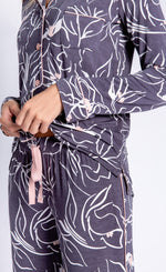 Load image into Gallery viewer, Front close up view of a woman wearing the pj salvage love lines set. This pj set is charcoal colored with white flowers drawn all over it and light pink detailing. The top has a button down front and long sleeves. The bottom is relaxed with a pink ribbon tie.
