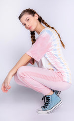 Load image into Gallery viewer, Front left side full body view of a woman crouching and wearing the PJ Salvage Sunset Hues short sleeve top and the pj salvage sunset hues banded pant. The top is white with yellow, blue, and pink tie dye on the sides. The bottoms are pink with crochet detailing on the sides and a banded hem. 

