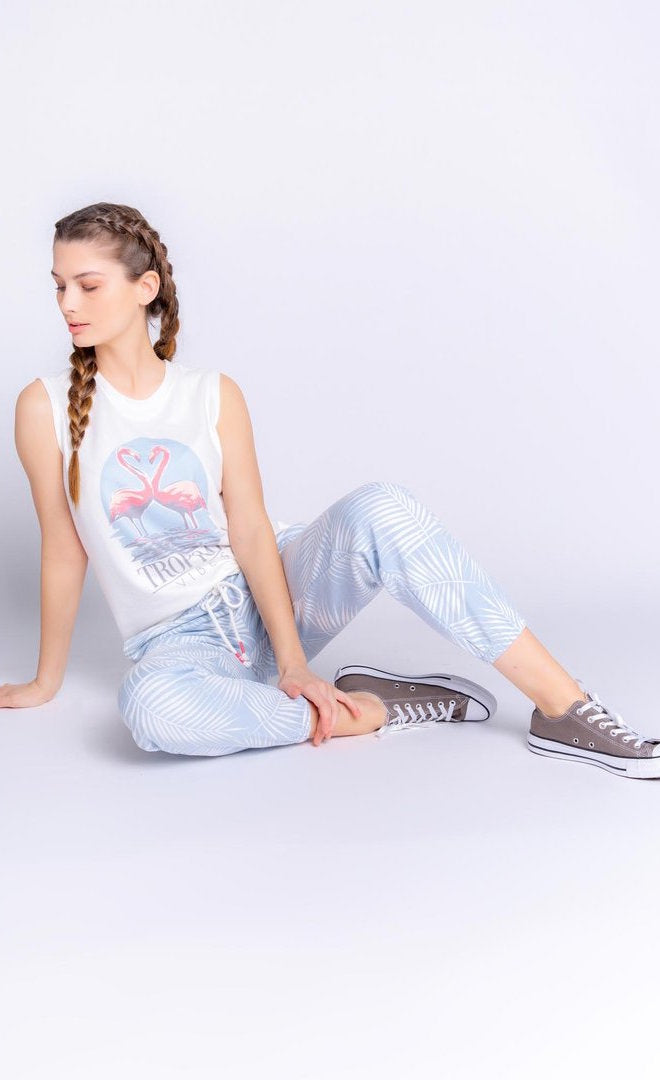 Front full body view of a woman sitting on the ground and wearing the pj salvage tropical vibes banded pant and the pj salvage tropical vibes tank. The tank is white with two flamingos printed on the front and the words "tropical vibes" underneath. The bottoms are sky blue with a white palm leaf print all over it.