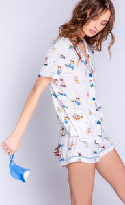 Front full body view of a woman wearing the pj salvage playful prints vacation mode pj set. This set includes a button down shirt with a shirt collar and pocket, shorts, and a sleep mask that says vacation mode on. The pj is ivory with a vacationing dog print all over it.
