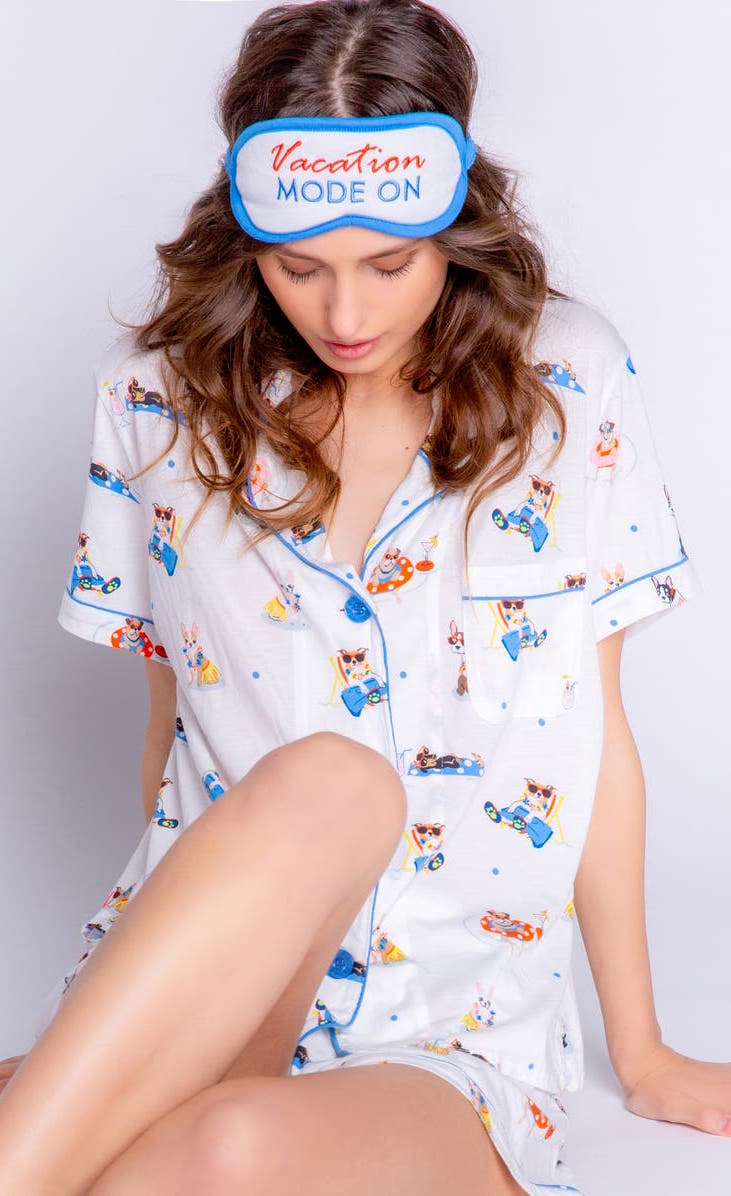 Front view of a woman sitting and wearing the pj salvage playful prints vacation mode pj set. This set includes a button down shirt with a shirt collar and pocket, shorts, and a sleep mask that says vacation mode on. The pj is ivory with a vacationing dog print all over it.