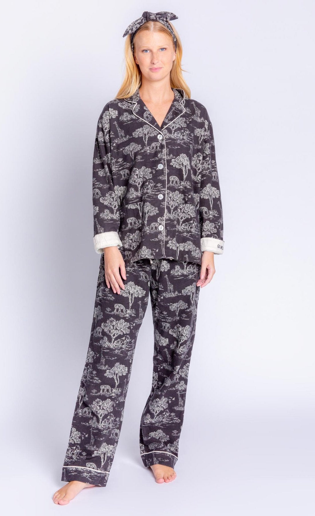 Front full body view of a woman wearing the PJ Salvage Go Wild PJ Set. This set is black with a white safari print all over it. It includes a long sleeve shirt and long pants. The cuffs are white and say go wild. The woman is also wearing a matching headband with a top knot.