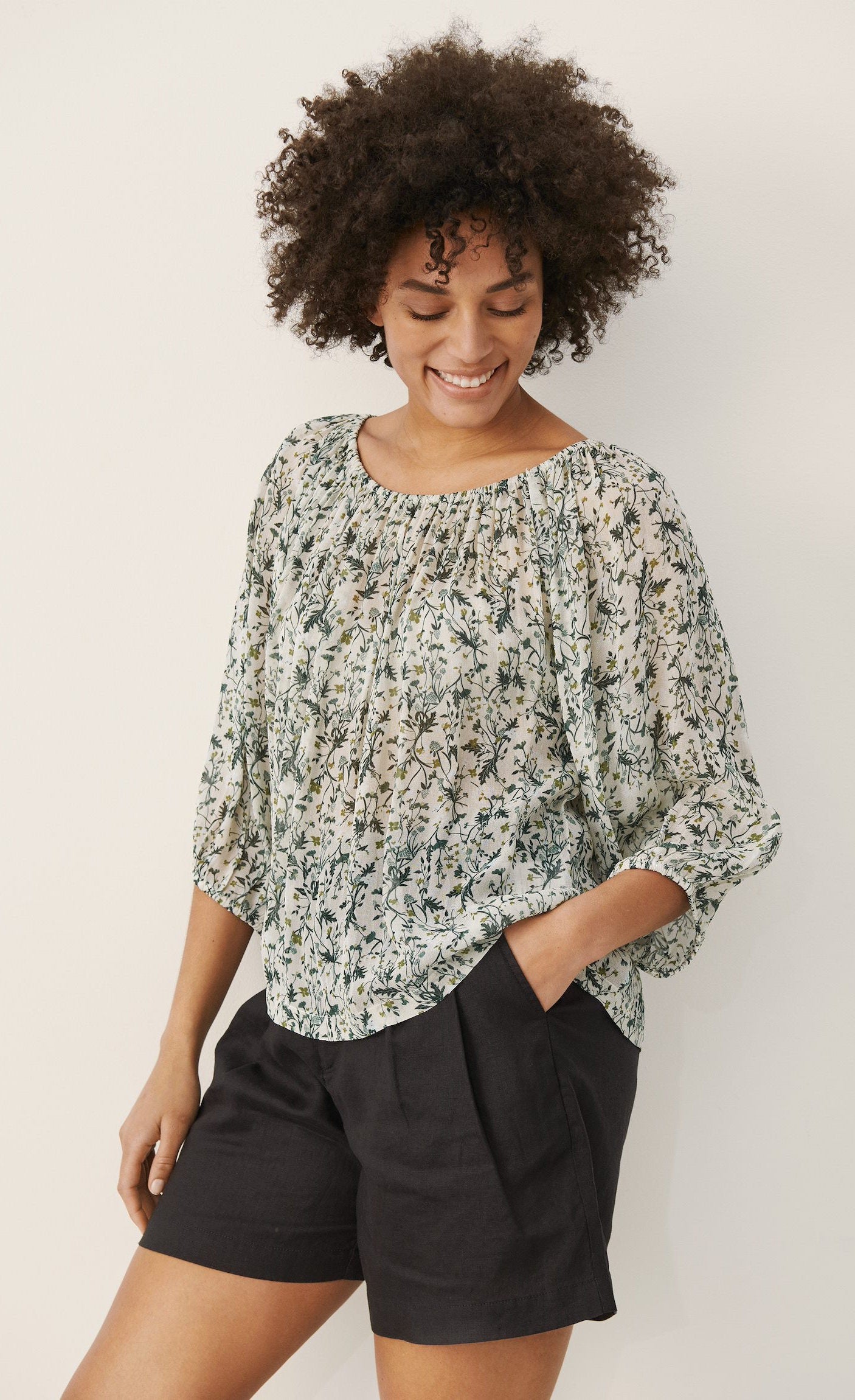 Front top half view of a woman wearing black shorts and the part two ingeborg floral top. This top has a small blue/green floral print, a gathered round neck, and 3/4 length wide sleeves.