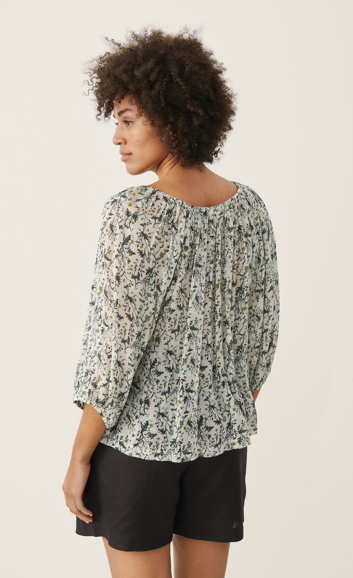 Back top half view of a woman wearing black shorts and the part two ingeborg floral top. This top has a small blue/green floral print, a gathered round neck, and 3/4 length wide sleeves.