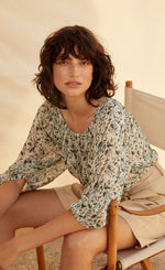 Load image into Gallery viewer, Front view of a woman sitting in a chair and wearing the part two ingeborg floral top. This top has a small blue/green floral print, a gathered round neck, and 3/4 length wide sleeves.
