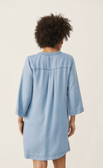 Load image into Gallery viewer, Back full body view of a woman wearing the part two ingeline dress. This dress is a light blue denim. It ends at the knees and has 3/4 length sleeves.
