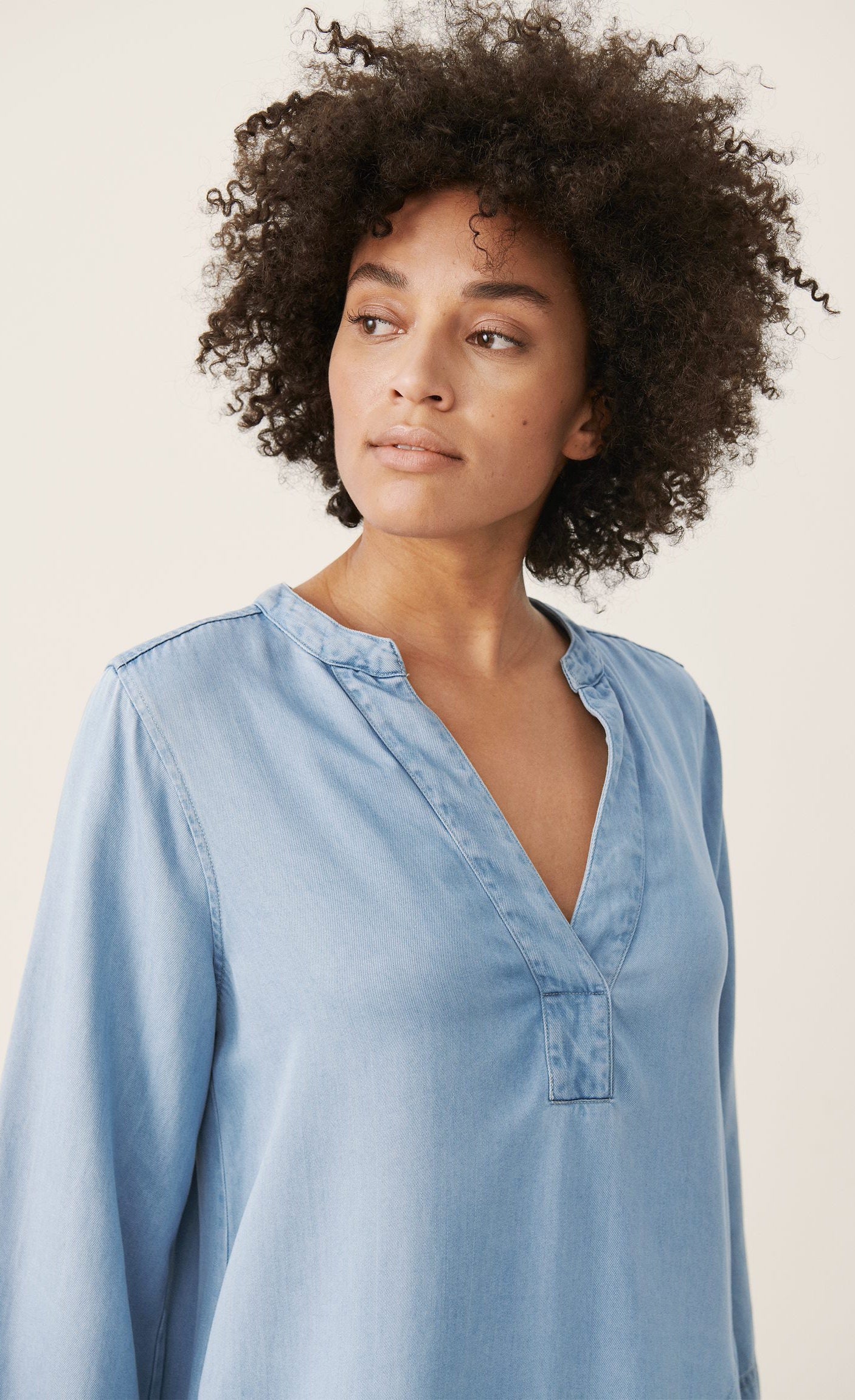 Front top half view of a woman wearing the part two ingeline dress. This dress is a light blue denim. It has 3/4 length sleeves and a v-neck. 