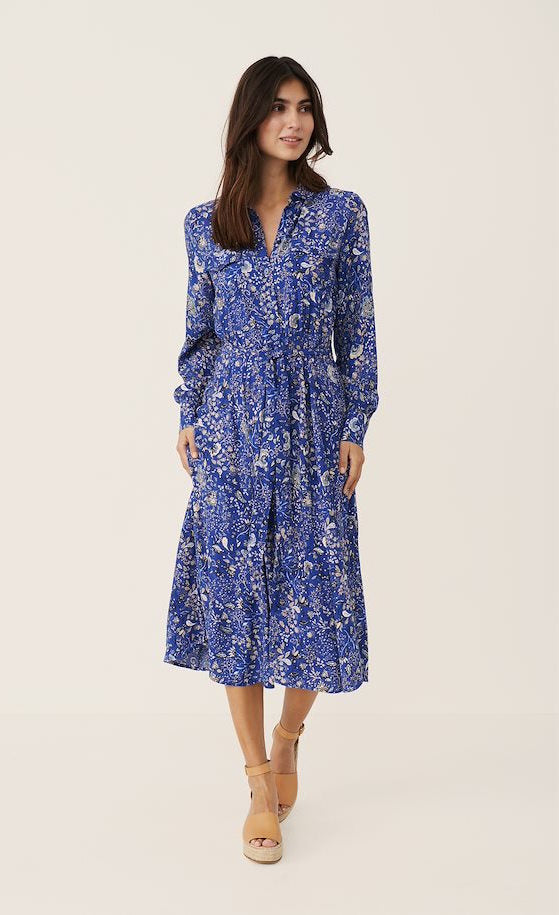 front full body view of a woman wearing the part two true paisley flower dress. This dress is ultramarine/vibrant blue color with white paisley floral print. The dress sits below the knees and has a button down front and tie belt.