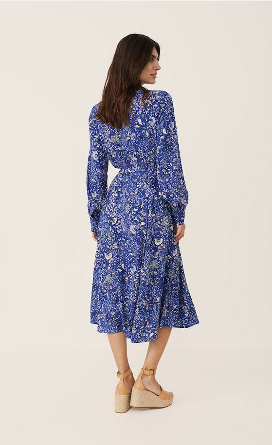 Back full body view of a woman wearing the part two true paisley flower dress. This dress is ultramarine/vibrant blue color with white paisley floral print. The dress sits below the knees and has a tie belt.