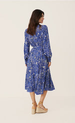 Load image into Gallery viewer, Back full body view of a woman wearing the part two true paisley flower dress. This dress is ultramarine/vibrant blue color with white paisley floral print. The dress sits below the knees and has a tie belt.
