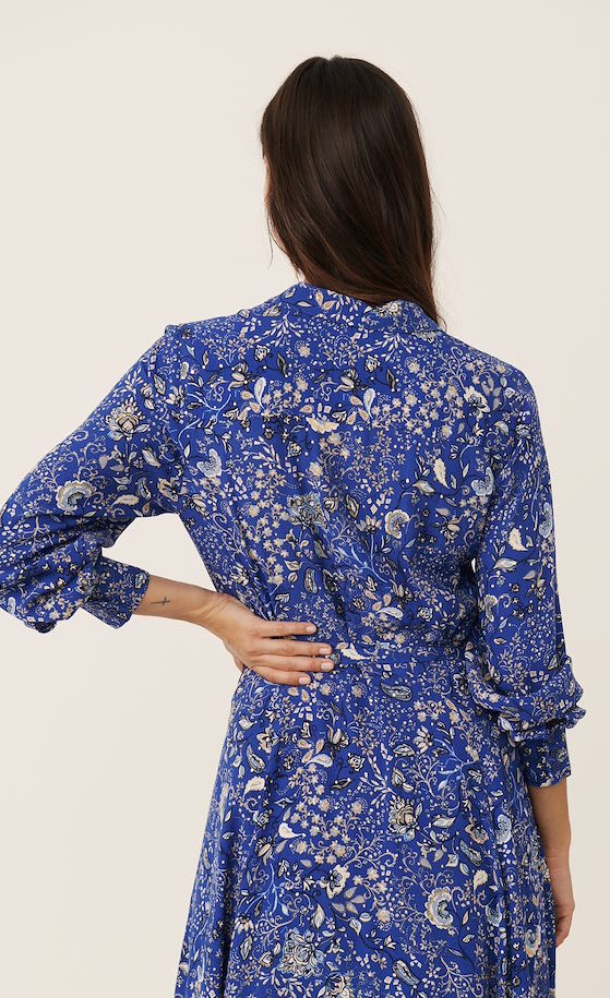 Back full body view of a woman wearing the part two true paisley flower dress. This dress is ultramarine/vibrant blue color with white paisley floral print. The dress has a tie belt at the waist.