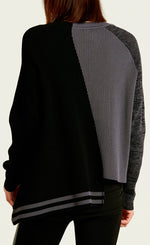 Load image into Gallery viewer, back top half view of a woman wearing black leather pants and the planet pima cotton 2 tone crewneck sweater. This sweater is black on the left side and grey on the right side. the left side also has a longer hem with grey striping, while the right side has a shorter hem with a washed black sleeve.
