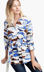 Load image into Gallery viewer, Front view of a woman leaning on a stool and wearing the Nic+Zoe In A Row Blouse. This blouse has a multi shaded blue and brown shibori print, 3/4 length sleeves, a button down front, and a front tie at the waist
