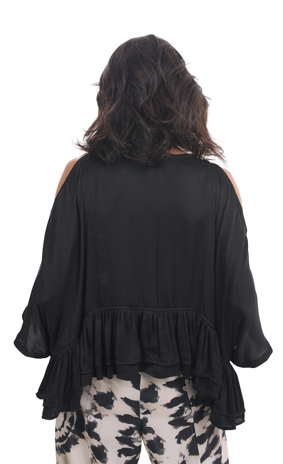 Back top half view of a woman wearing the rhys ruffle blouse in black. This top has a cold shoulder, 3/4 length sleeves, and a ruffled hem. On the bottom the woman is wearing black and white printed pants.