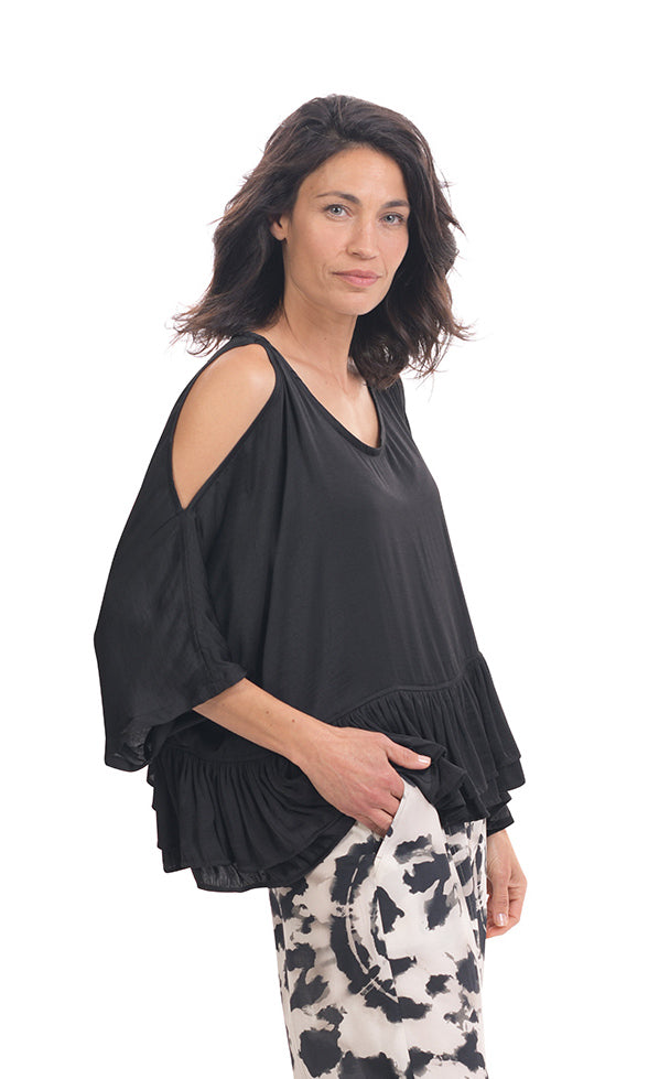 Right side top half view of a woman wearing the rhys ruffle blouse in black. This top has a cold shoulder, a round neck, 3/4 length sleeves and a ruffled hem. On the bottom the woman is wearing black and white printed pants.
