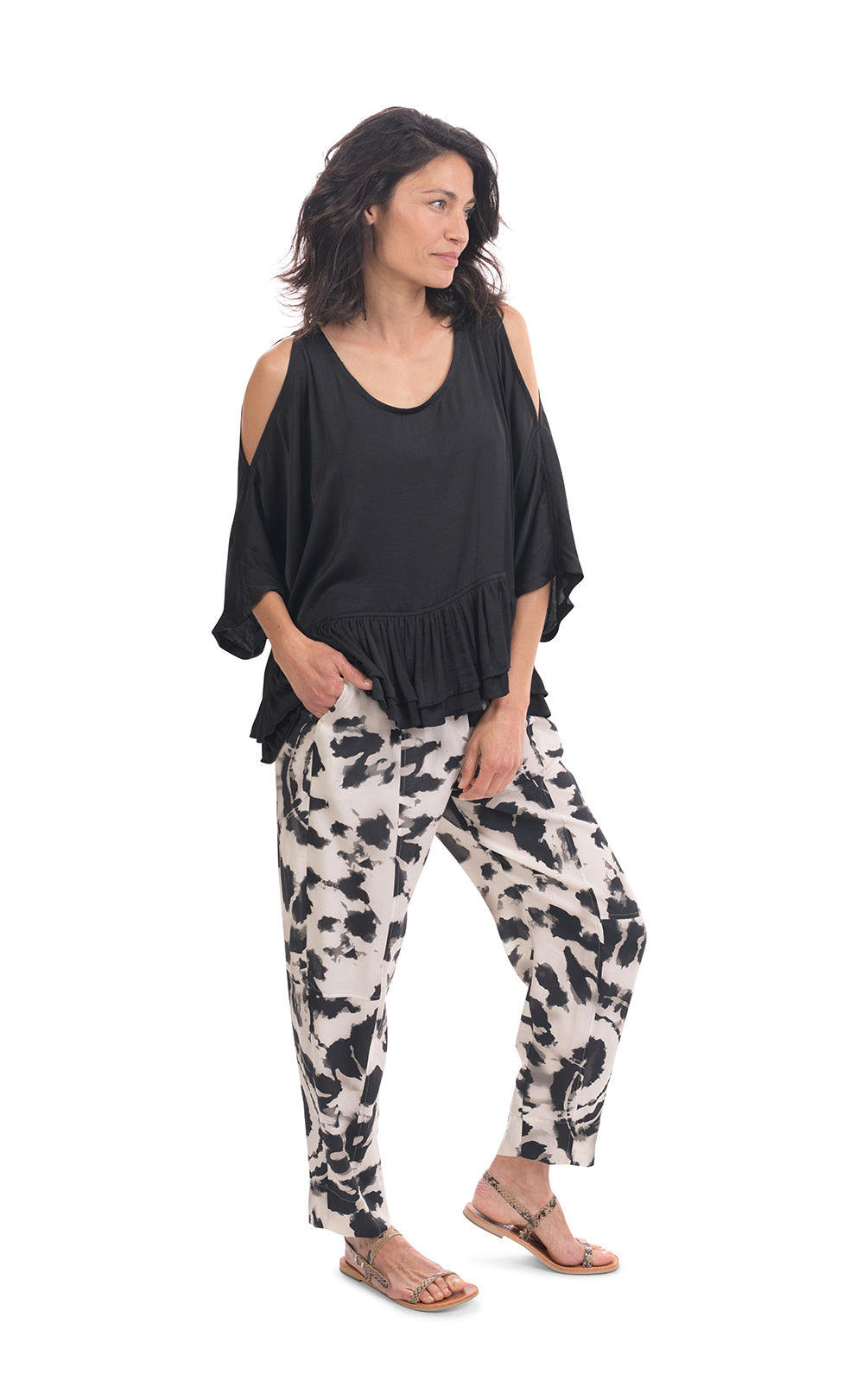Front full body view of a woman wearing the rhys ruffle blouse in black. This top has a cold shoulder, a round neck, 3/4 length sleeves and a ruffled hem. On the bottom the woman is wearing black and white printed pants.