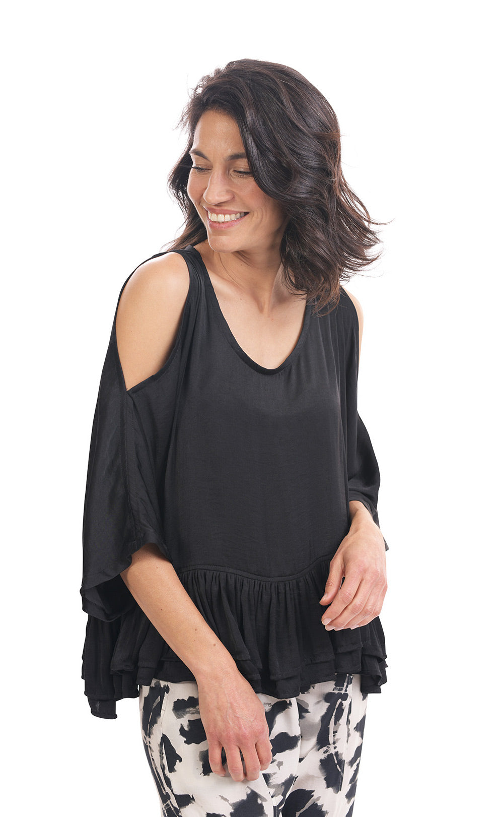 Front top half view of a woman wearing the rhys ruffle blouse in black. This top has a cold shoulder, a round neck, 3/4 length sleeves and a ruffled hem. On the bottom the woman is wearing black and white printed pants.