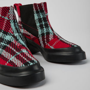 Close up front view of a pair of the camper blanket/rain multicolor boot. These boots have a chelsea style with elastic sides. The boots have a red, black, and blue plaid print and a chunky black heel.
