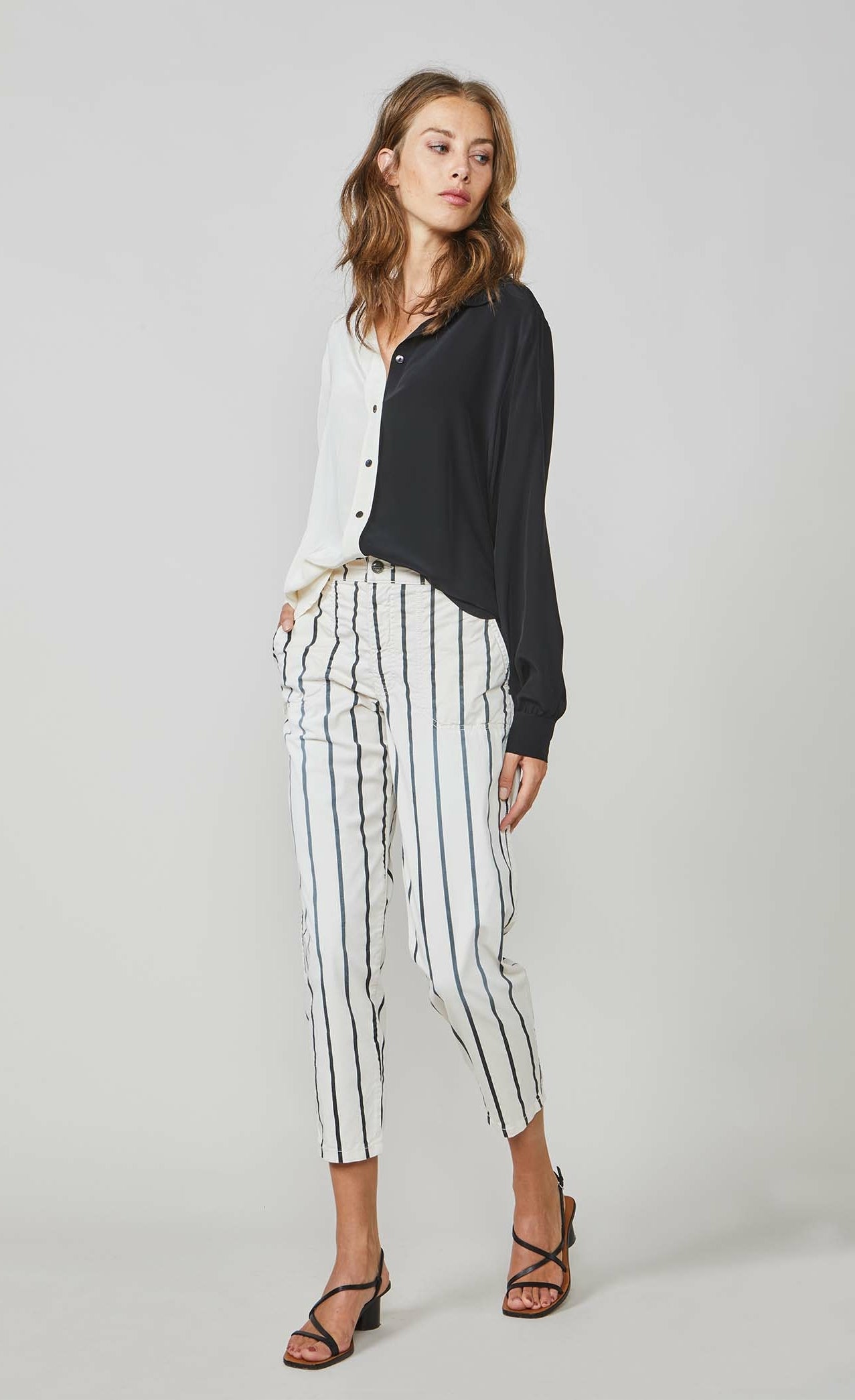Front full body view of a woman wearing the summum color block blouse. This blouse is black on the left side and ivory on the right side. It has a button up front and long sleeves. On the bottom the model is wearing the summum striped trouser
