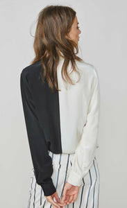 Back top half view of a woman wearing the summum color block blouse. This blouse is black on the left side and ivory on the right side. It has long sleeves with wide cuffs.