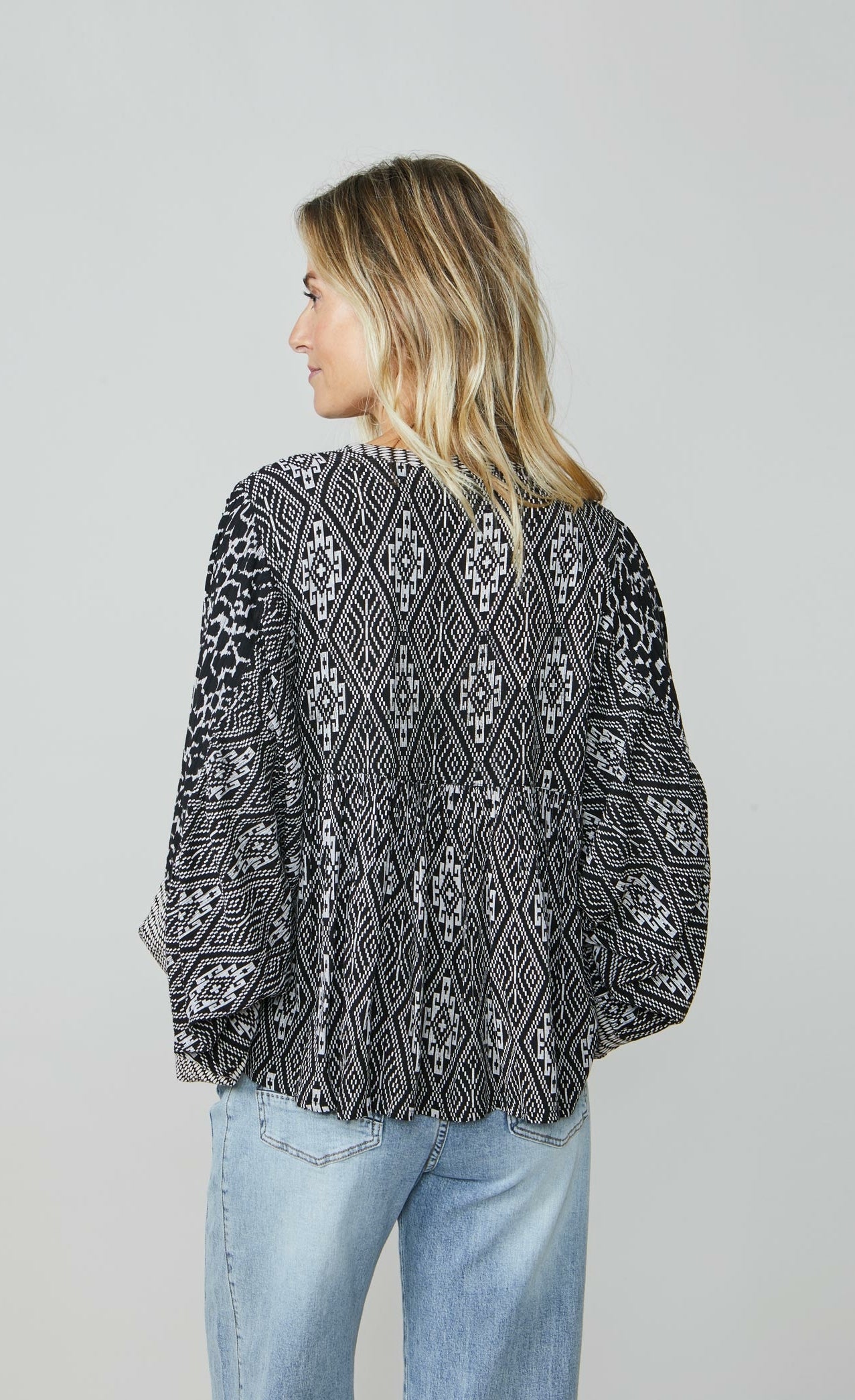 Back top half view of a woman wearing wide leg jeans and the summum kimono printed top. This top is black with a white print. It has an oversized silhouette and long puffy sleeves.