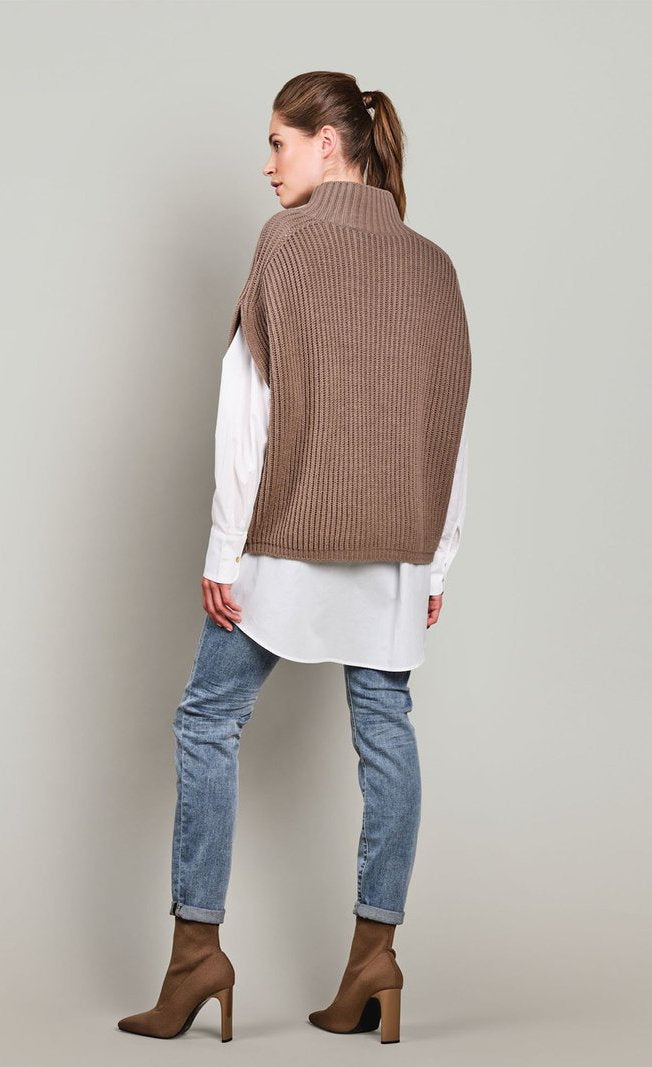 Back full body view of a woman wearing jeans, a white long sleeve shirt, and the summum sleeveless cable knit sweater. This sweater is ribbed with a stand collar.