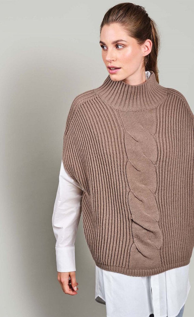 Front top half view of a woman wearing jeans, a white long sleeve shirt, and the summum sleeveless cable knit sweater. This sweater is ribbed with a cable knit band down the middle front. The sweater also has a ribbed stand collar.