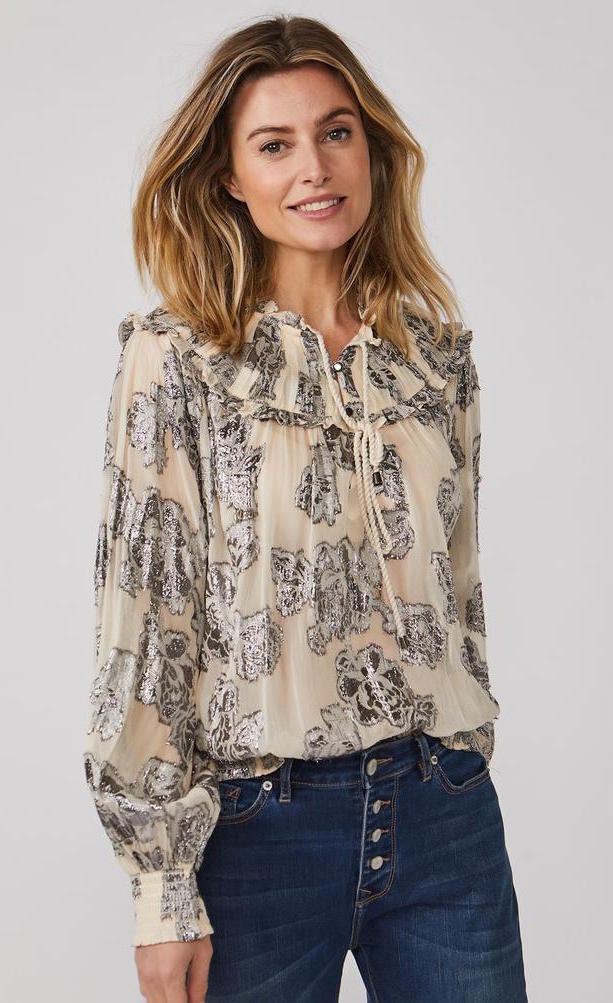 Front top half view of a woman wearing the summum lurex flower blouse. This blouse is cream/shell colored with metallic flowers all over it, a tie neck, balloon sleeves, and a round shoulder panel with ruffles.