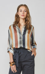 Load image into Gallery viewer, Front full body view of a woman wearing full navy capris and the summum multicolor striped blouse. This blouse has rust, mustard, blue, and green stripes separated by a light blue. The blouse also has a button up front and rolled up long sleeves.
