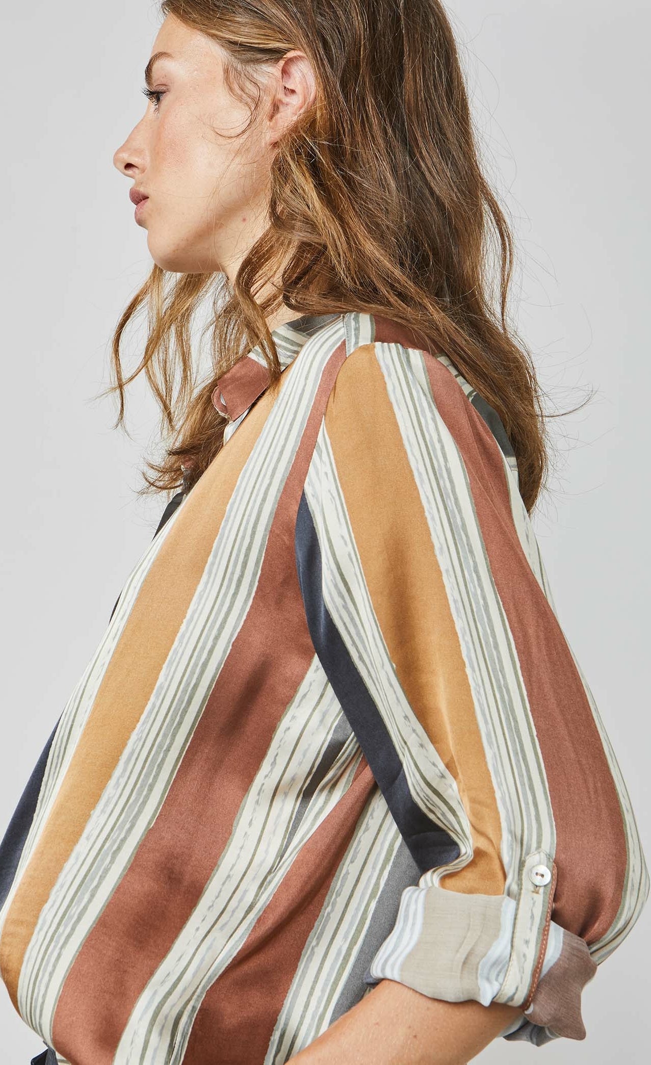 Side close up view of a woman wearing the summum multicolor striped blouse. This blouse has rust, mustard, blue, and green stripes separated by a light blue. The blouse also has a button up front and rolled up long sleeves.