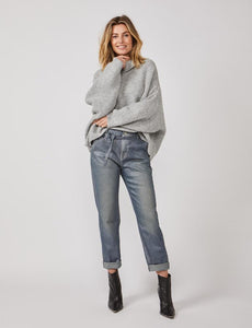 Front full body view of a woman wearing a grey sweater and the summum straight blue daze denim pants. These jeans are blue-grey with a shimmer. They have a cropped and cuffed bottom, pockets, and a tie belt.