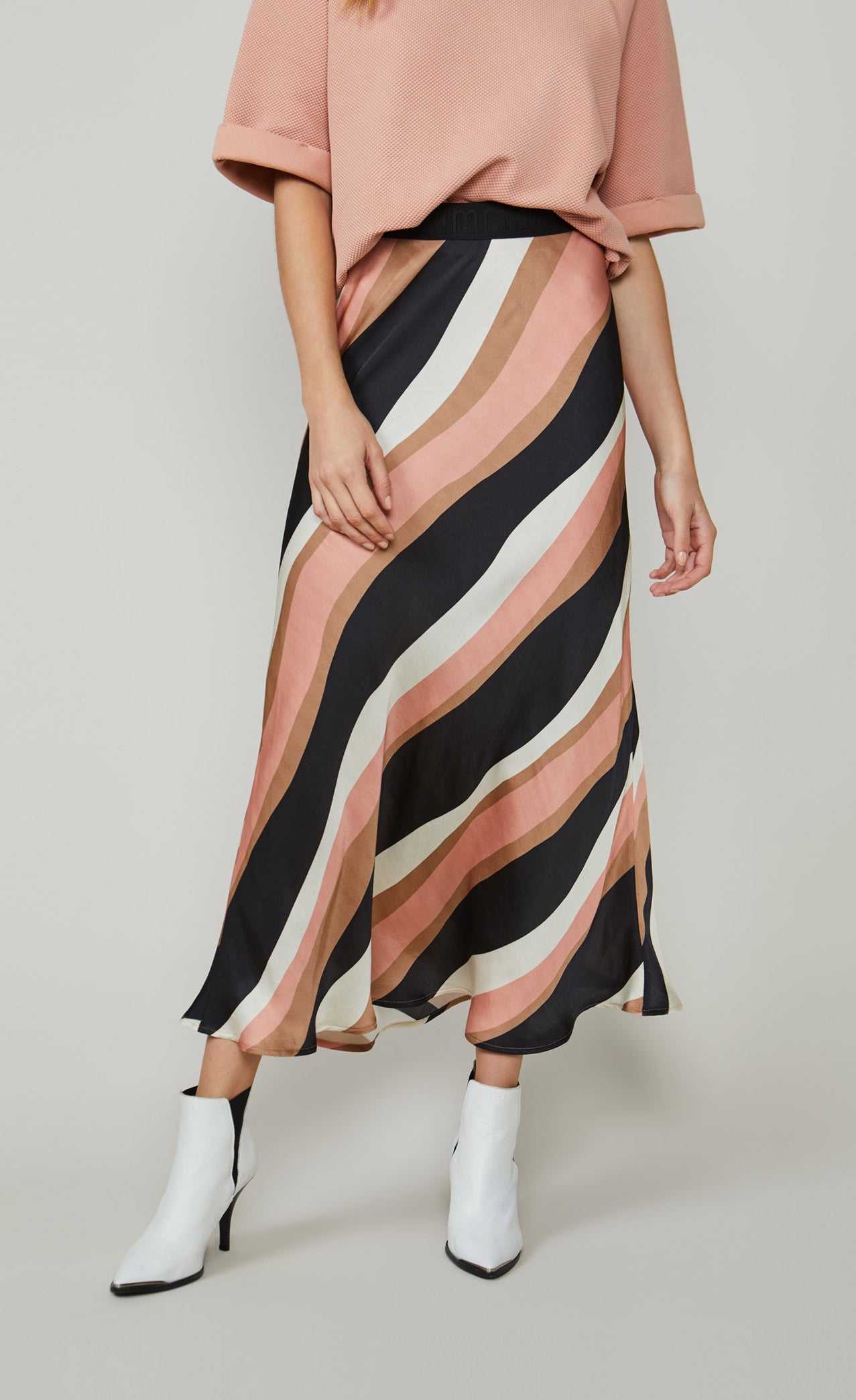 Front bottom half view of a woman wearing a pink top and the summum striped waves printed maxi skirt. This skirt has different sized wavy stripes in black pink, taupe, and white that run diagonally across it. The skirt ends just above the ankles.