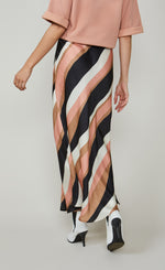 Load image into Gallery viewer, Back bottom half view of a woman wearing a pink top and the summum striped waves printed maxi skirt. This skirt has different sized wavy stripes in black pink, taupe, and white that run diagonally across it. The skirt ends just above the ankles.
