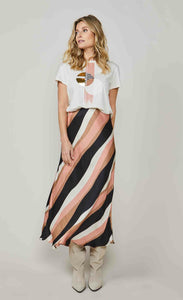 Front full body view of a woman wearing a white t-shirt and the summum striped waves printed maxi skirt. This skirt has different sized wavy stripes in black pink, taupe, and white that run diagonally across it. The skirt ends just above the ankles.