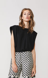Front top half view of a woman wearing the summum shoulder pad top. This top is sleeves and black with a v-like shape. On the bottom the woman is wearing a white pant with black print.