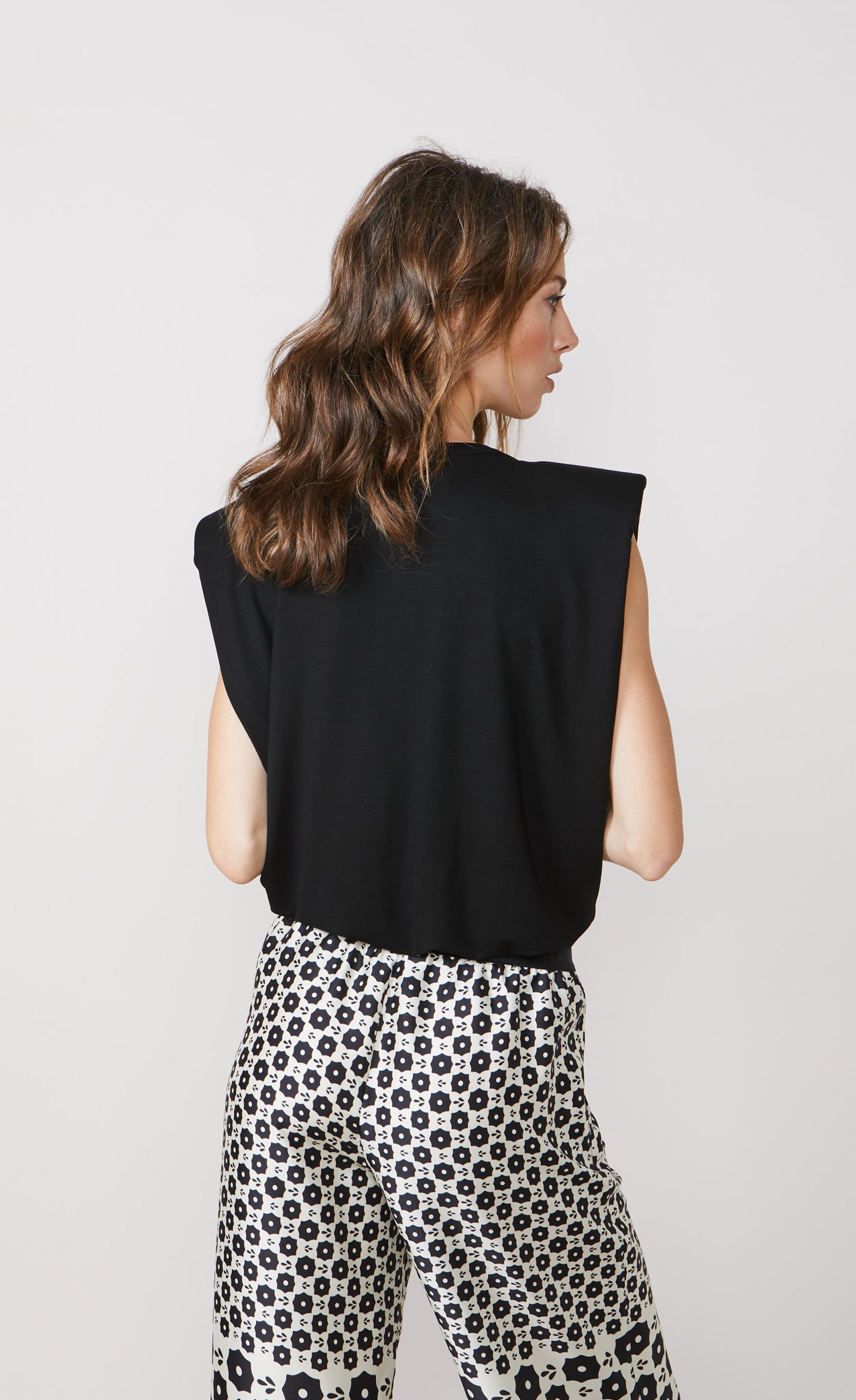 Back top half view of a woman wearing the summum shoulder pad top. This top is sleeves and black with a v-like shape. On the bottom the woman is wearing a white pant with black print.