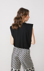 Load image into Gallery viewer, Back top half view of a woman wearing the summum shoulder pad top. This top is sleeves and black with a v-like shape. On the bottom the woman is wearing a white pant with black print.
