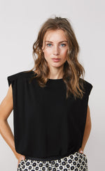 Load image into Gallery viewer, Front top half view of a woman wearing the summum shoulder pad top. This top is sleeves and black with a v-like shape. On the bottom the woman is wearing a white pant with black print.
