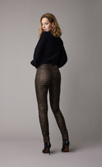 Load image into Gallery viewer, Back full body view of a woman wearing a black top and the summum Skinny trousers with foil print. This pant is caramel bronze colored with a small metallic gold and brown leaf print all over it.
